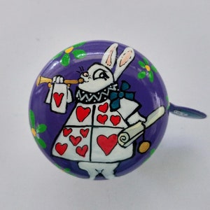 Puede incluir: Campana de bicicleta morada con un diseño pintado a mano de un conejo blanco que lleva un disfraz de carta de juego y sostiene una trompeta. El conejo tiene corazones rojos en su disfraz y una pajarita azul.