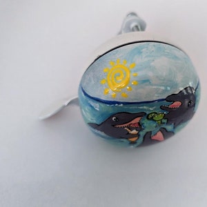 Puede incluir: Una campanilla de bicicleta blanca con un dise&ntilde;o pintado de tres delfines nadando en el oc&eacute;ano con un sol amarillo en el cielo.