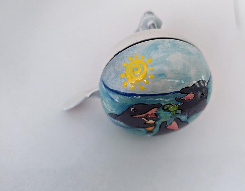 Puede incluir: Una campanilla de bicicleta blanca con un dise&ntilde;o pintado de tres delfines nadando en el oc&eacute;ano con un sol amarillo en el cielo.