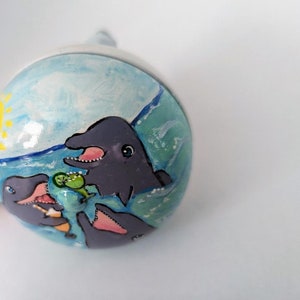 Puede incluir: Un adorno de cer&aacute;mica pintado a mano con tres delfines nadando en agua azul con un sol amarillo en el fondo. Los delfines est&aacute;n pintados en tonos grises y tienen dientes y ojos blancos.