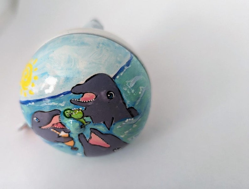 Puede incluir: Un adorno de cer&aacute;mica pintado a mano con tres delfines nadando en agua azul con un sol amarillo en el fondo. Los delfines est&aacute;n pintados en tonos grises y tienen dientes y ojos blancos.