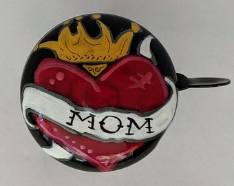 May include: Black bicycle bell with a hand-painted design of a red heart with a gold crown on top and a white banner with the word "MOM" on it.