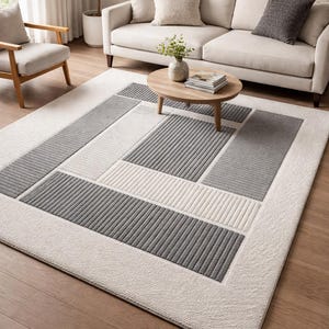Peut inclure: Un tapis rectangulaire avec un motif géométrique dans les tons de gris et de blanc. Le tapis présente une bordure texturée et un motif central de rayures et de blocs alternés. Il est posé sur un parquet.