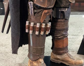 May include: A detailed view of a costume's lower half, featuring brown leather boots and leg armor. The armor includes multiple silver cylinders held by leather straps. The costume has a weathered appearance, suggesting use in a rugged environment.
