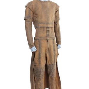 May include: A tan-colored costume displayed on a mannequin. The outfit includes a long-sleeved top with shoulder pads and a matching pair of pants. The pants have knee patches and a long, split skirt. The costume has a distressed, worn appearance.