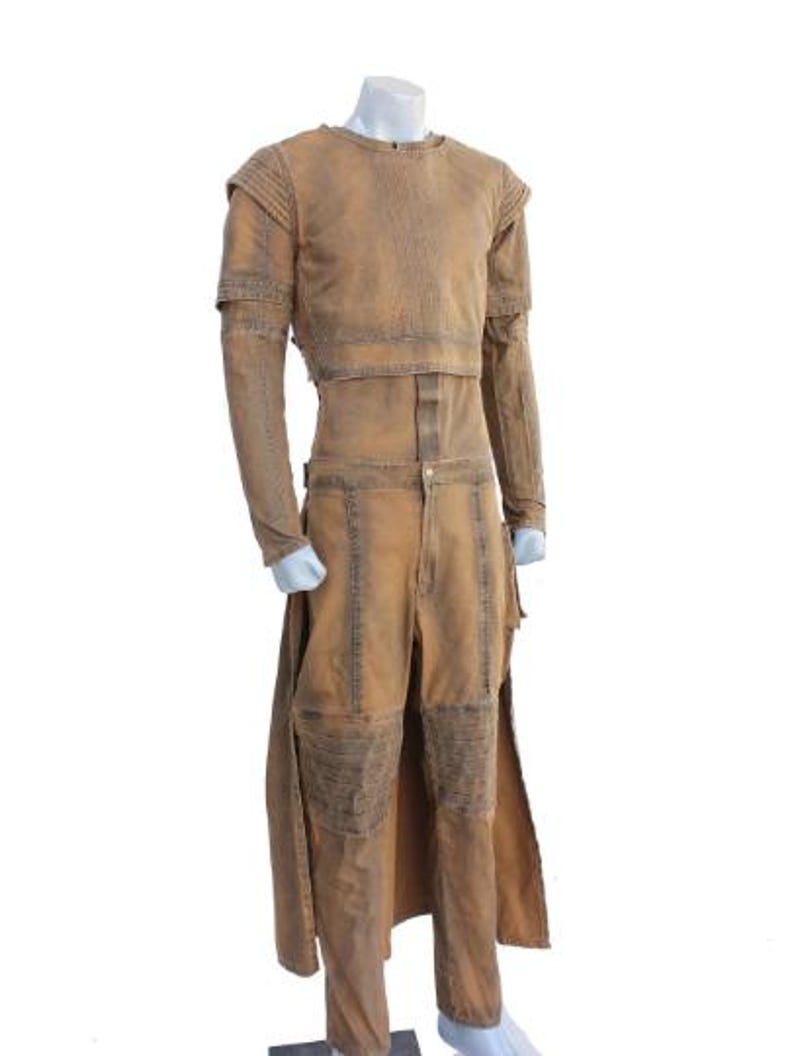 May include: A tan-colored costume displayed on a mannequin. The outfit includes a long-sleeved top with shoulder pads and a matching pair of pants. The pants have knee patches and a long, split skirt. The costume has a distressed, worn appearance.