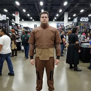 May include: A person wearing a brown costume with a high collar, geometric patterns, and black boots. The outfit includes a top, pants, and a belt-like detail. The background shows a convention setting with various booths and people.
