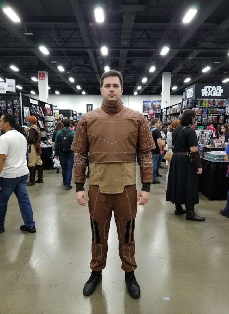 May include: A person wearing a brown costume with a high collar, geometric patterns, and black boots. The outfit includes a top, pants, and a belt-like detail. The background shows a convention setting with various booths and people.