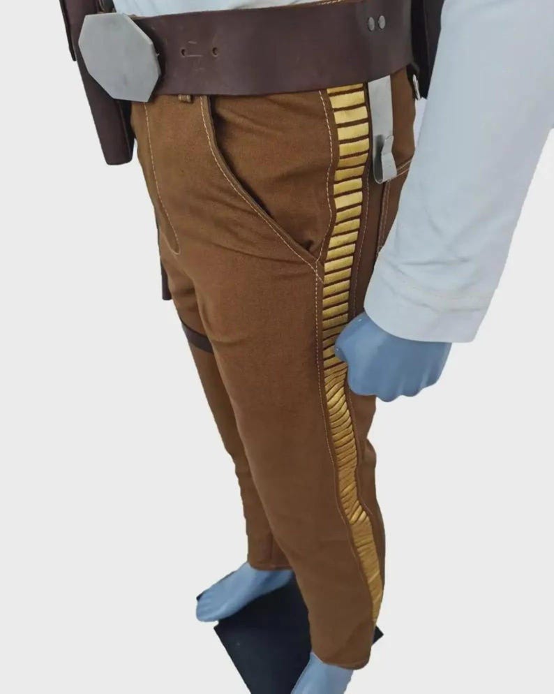 Pode incluir: Cal&ccedil;as castanhas com uma faixa decorativa vertical de cor dourada ao longo da perna exterior, combinadas com um cinto castanho largo que inclui uma grande fivela met&aacute;lica hexagonal e um coldre, usadas com uma camisa branca de manga comprida.