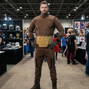 May include: A person wearing a brown and tan costume with a high collar, gloves, and boots. The costume has a quilted pattern on the sleeves and legs. The person is standing in a convention center with various booths and displays in the background.