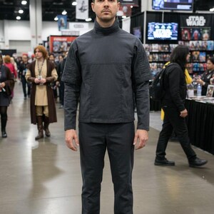K&ouml;nnte beinhalten: Eine Person in einem dunkelgrauen Kost&uuml;m, bestehend aus einem lang&auml;rmeligen Oberteil mit hohem Kragen und passender Hose. Das Outfit ist wahrscheinlich ein Cosplay oder Kost&uuml;m, m&ouml;glicherweise inspiriert von einem Science-Fiction-Thema.