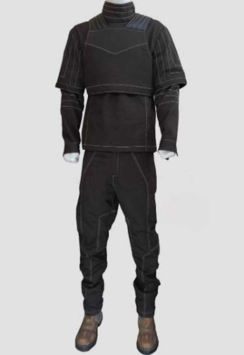 Pu&ograve; includere: Un completo tattico marrone scuro con dettagli di cuciture bianche. L'outfit include una giacca a collo alto con dettagli sulle spalle e un pannello sul petto, maniche lunghe e pantaloni abbinati. Il completo &egrave; esposto su un manichino con stivali marroni.