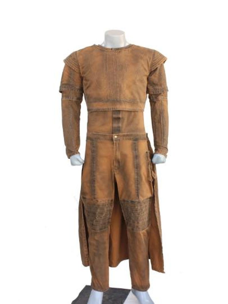 May include: A two-piece costume in shades of brown, displayed on a mannequin. The top has long sleeves and a structured design, while the pants feature a split front and knee patches. The outfit has a distressed, textured appearance.