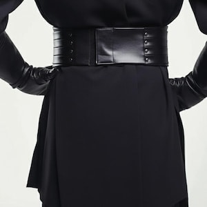 May include: A wide, black leather belt with a rectangular buckle and rivet detailing. The belt is worn over a black garment, creating a defined waistline. The overall aesthetic is sleek and modern, suitable for fashion-forward outfits.