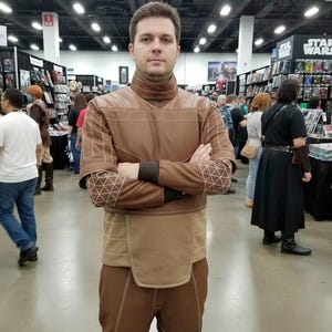 May include: A person wearing a brown costume with a high collar and geometric patterns on the sleeves. The costume has a tan panel at the waist and is worn in a convention setting with other people and merchandise in the background.