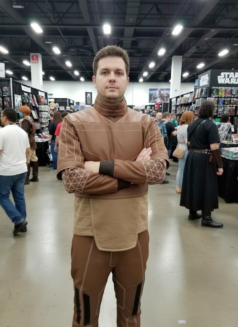 May include: A person wearing a brown costume with a high collar and geometric patterns on the sleeves. The costume has a tan panel at the waist and is worn in a convention setting with other people and merchandise in the background.