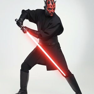 May include: A figure in a black outfit with a red and black face, wielding a red lightsaber. The costume comprises a black tunic, trousers, boots, and gloves. The lightsaber is illuminated, and the figure is in a combat pose.