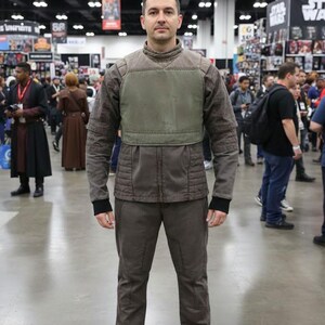 Puede incluir: Una persona con un disfraz marr&oacute;n y verde oliva. El atuendo incluye una chaqueta con cuello alto y peto, pantalones a juego y zapatos negros. El conjunto parece ser un cosplay o disfraz.