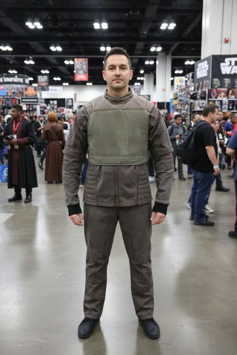 Puede incluir: Una persona con un disfraz marr&oacute;n y verde oliva. El atuendo incluye una chaqueta con cuello alto y peto, pantalones a juego y zapatos negros. El conjunto parece ser un cosplay o disfraz.