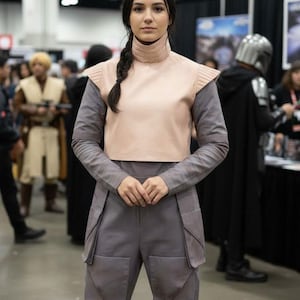May include: A person wearing a cosplay costume. The outfit includes a light pink top with shoulder pads, a gray long-sleeved shirt, and gray pants with large pockets. The person has dark hair in a braid.