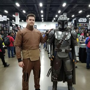 May include: A person in a brown costume stands next to a Mandalorian costume. The Mandalorian armor is silver and gray, with a helmet and cape. The person in the brown costume has a high-collared top and matching pants. The background shows a convention setting.