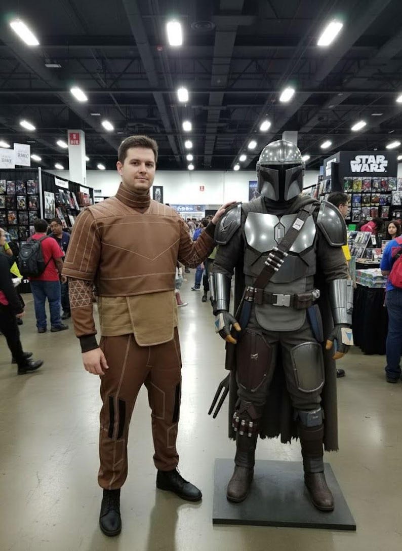 May include: A person in a brown costume stands next to a Mandalorian costume. The Mandalorian armor is silver and gray, with a helmet and cape. The person in the brown costume has a high-collared top and matching pants. The background shows a convention setting.