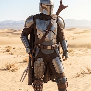May include: A person in a silver helmet and armor, standing in a desert landscape. The armor includes shoulder pads, chest plate, and leg guards. A brown cape and utility belt are also visible. A weapon is held on the right shoulder.