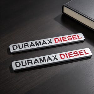 Può includere: Due emblemi rettangolari in metallo color argento con la scritta DURAMAX in nero e DIESEL in rosso. Gli emblemi presentano una finitura metallica liscia con caratteri in rilievo, progettati per l'applicazione sulla carrozzeria di veicoli.