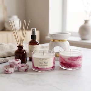 Puede incluir: Una colección de artículos de fragancia para el hogar que incluye dos velas en frasco de vidrio con cera veteada en blanco y rosa, etiquetadas Purewick Rose Kiss, un difusor de varillas de vidrio ámbar, un atomizador y tres ceras aromáticas.