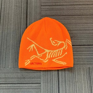 May include: Bright orange beanie with a cream-colored graphic and the text "ARC'TERYX" in red. The hat is made of a knitted material and is designed to keep the head warm. The beanie is a winter accessory.