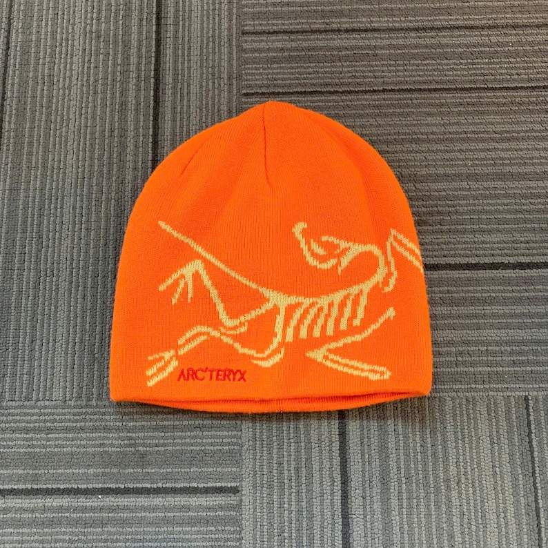 May include: Bright orange beanie with a cream-colored graphic and the text "ARC'TERYX" in red. The hat is made of a knitted material and is designed to keep the head warm. The beanie is a winter accessory.