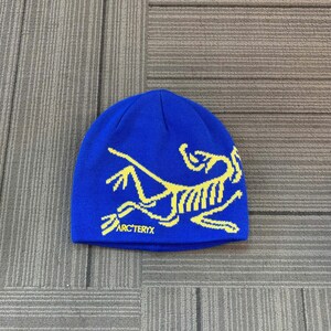 May include: A blue beanie with a yellow graphic design and the text "ARC'TERYX" at the bottom. The beanie is made of a soft, knitted material and is suitable for cold weather.