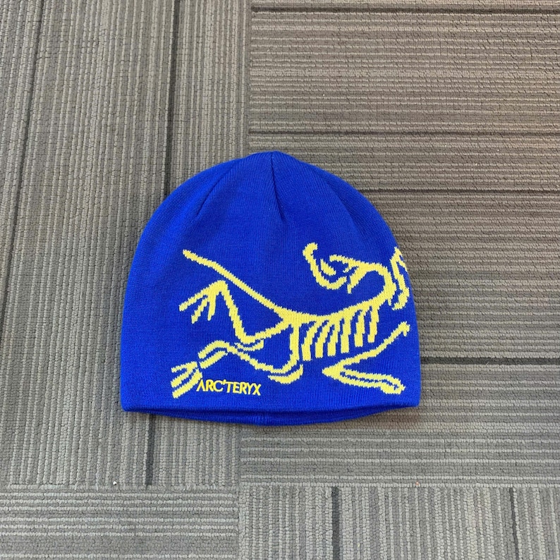 May include: A blue beanie with a yellow graphic design and the text "ARC'TERYX" at the bottom. The beanie is made of a soft, knitted material and is suitable for cold weather.