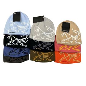May include: A stack of beanies in various colors, including light blue, gray, black, and orange. Each beanie features a white graphic and the brand name "Arc'teryx". The beanies have a folded brim and a tag attached.