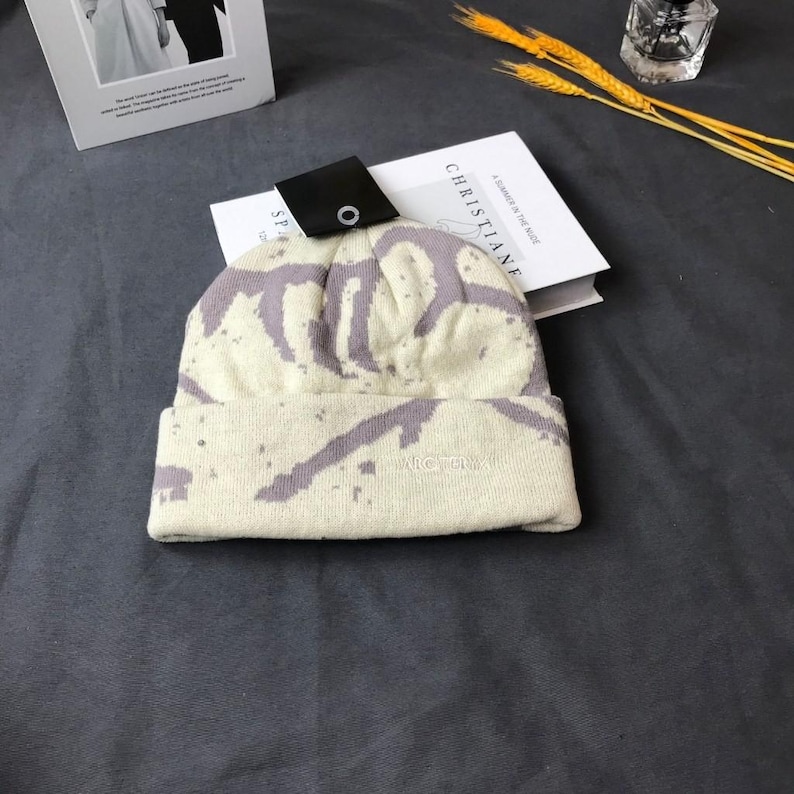 Peut inclure: Un bonnet cr&egrave;me avec un motif &eacute;clabouss&eacute; lavande. Le bord repli&eacute; porte le mot "VICTORY" en petites lettres brod&eacute;es. Une &eacute;tiquette noire avec un bouton argent&eacute; est attach&eacute;e. Un livre avec le mot "CHRISTIANE" est visible.
