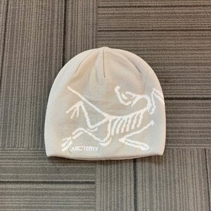 May include: A light beige beanie featuring a white graphic of a stylized animal skeleton. The brand name "ARC'TERYX" is printed in white below the graphic. The hat is made of a soft, knitted material.
