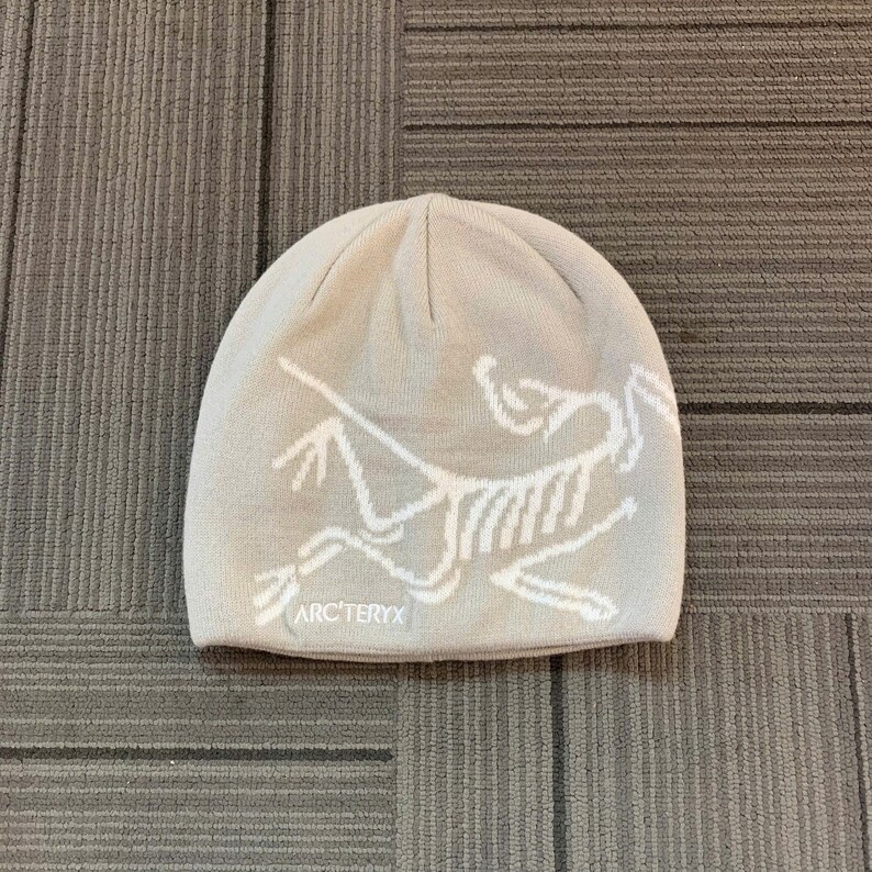 May include: A light beige beanie featuring a white graphic of a stylized animal skeleton. The brand name "ARC'TERYX" is printed in white below the graphic. The hat is made of a soft, knitted material.