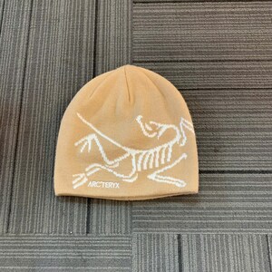 May include: A tan beanie with a white graphic design and the text "ARC'TERYX" at the bottom. The hat is made of a knitted material and is likely designed for cold weather.