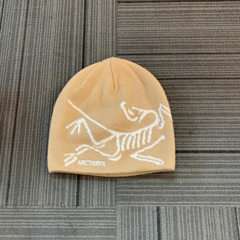 May include: A tan beanie with a white graphic design and the text "ARC'TERYX" at the bottom. The hat is made of a knitted material and is likely designed for cold weather.