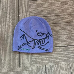 May include: A periwinkle blue beanie with a black graphic design and the word "Arc'teryx" in black text. The beanie is made of a soft, knitted material. This hat is suitable for outdoor activities.
