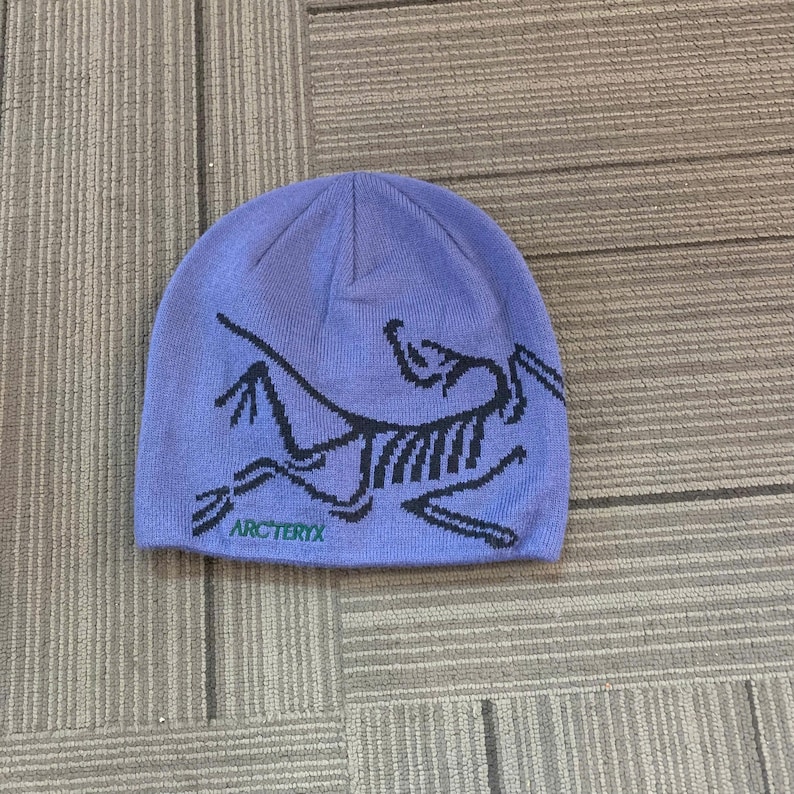 May include: A periwinkle blue beanie with a black graphic design and the word "Arc'teryx" in black text. The beanie is made of a soft, knitted material. This hat is suitable for outdoor activities.