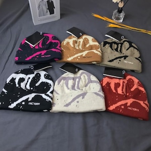 Peut inclure: Six bonnets de couleurs vari&eacute;es, dont noir, marron et rouge, chacun avec un motif &eacute;clabouss&eacute; unique. Les bonnets ont une &eacute;tiquette attach&eacute;e et le nom de la marque "ARC'TERYX" est visible &agrave; l'avant.