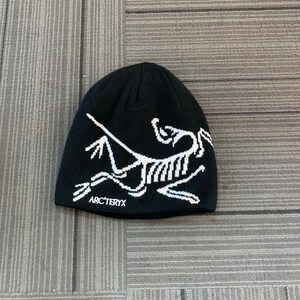 May include: Black beanie with a white graphic of a stylized skeleton and the brand name "ARC'TERYX" in white. The hat is made of a knit material and is laying on a gray surface.