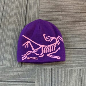 May include: A purple beanie with a pink graphic design and the text "ARC'TERYX" at the bottom. The hat is made of a soft material and is likely designed for cold weather.
