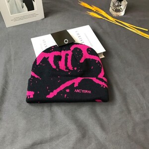 Peut inclure: Bonnet noir avec un motif abstrait rose vif. Le chapeau porte le nom de la marque "ARC'TERYX" en rose. Le bonnet est pli&eacute; au bord et repose sur un livre blanc.