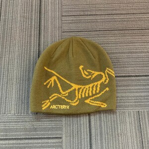 May include: An olive green beanie with a yellow graphic of a stylized animal and the text "ARC'TERYX". The hat is made of a knitted material and is designed to keep the head warm.