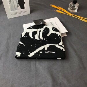 Peut inclure: Bonnet noir et blanc avec un motif unique. Le bonnet porte le nom de la marque "ARC'TERYX" en lettres blanches. Le chapeau est pli&eacute; et une &eacute;tiquette est attach&eacute;e.