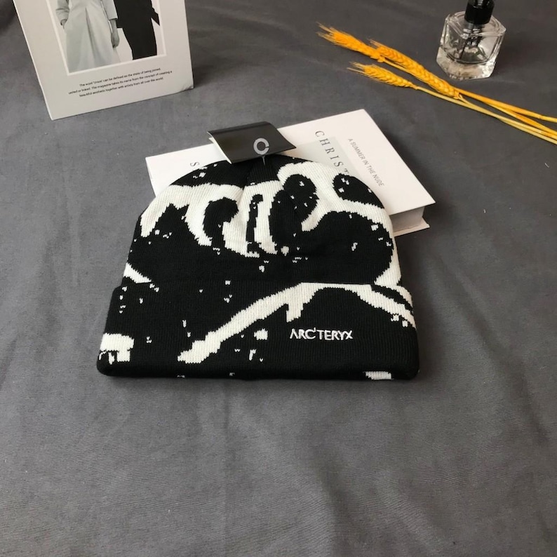Peut inclure: Bonnet noir et blanc avec un motif unique. Le bonnet porte le nom de la marque "ARC'TERYX" en lettres blanches. Le chapeau est pli&eacute; et une &eacute;tiquette est attach&eacute;e.