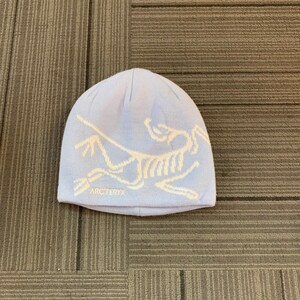 May include: A light blue beanie with a white graphic design and the text "ARC'TERYX" at the bottom. The beanie is made of a soft, knitted material and is designed to keep the head warm.