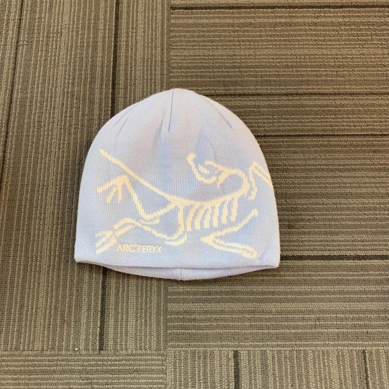 May include: A light blue beanie with a white graphic design and the text "ARC'TERYX" at the bottom. The beanie is made of a soft, knitted material and is designed to keep the head warm.